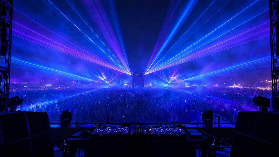 Widok zza stanowiska DJ-a na wielkiej scenie: niebieskie i fioletowe lasery nad tłumem